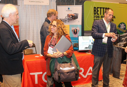 Attendees engage in conversation at the TOMRA Sorting Inc. booth, where a woman with curly hair holds a laptop and bag while speaking with others at the conference.