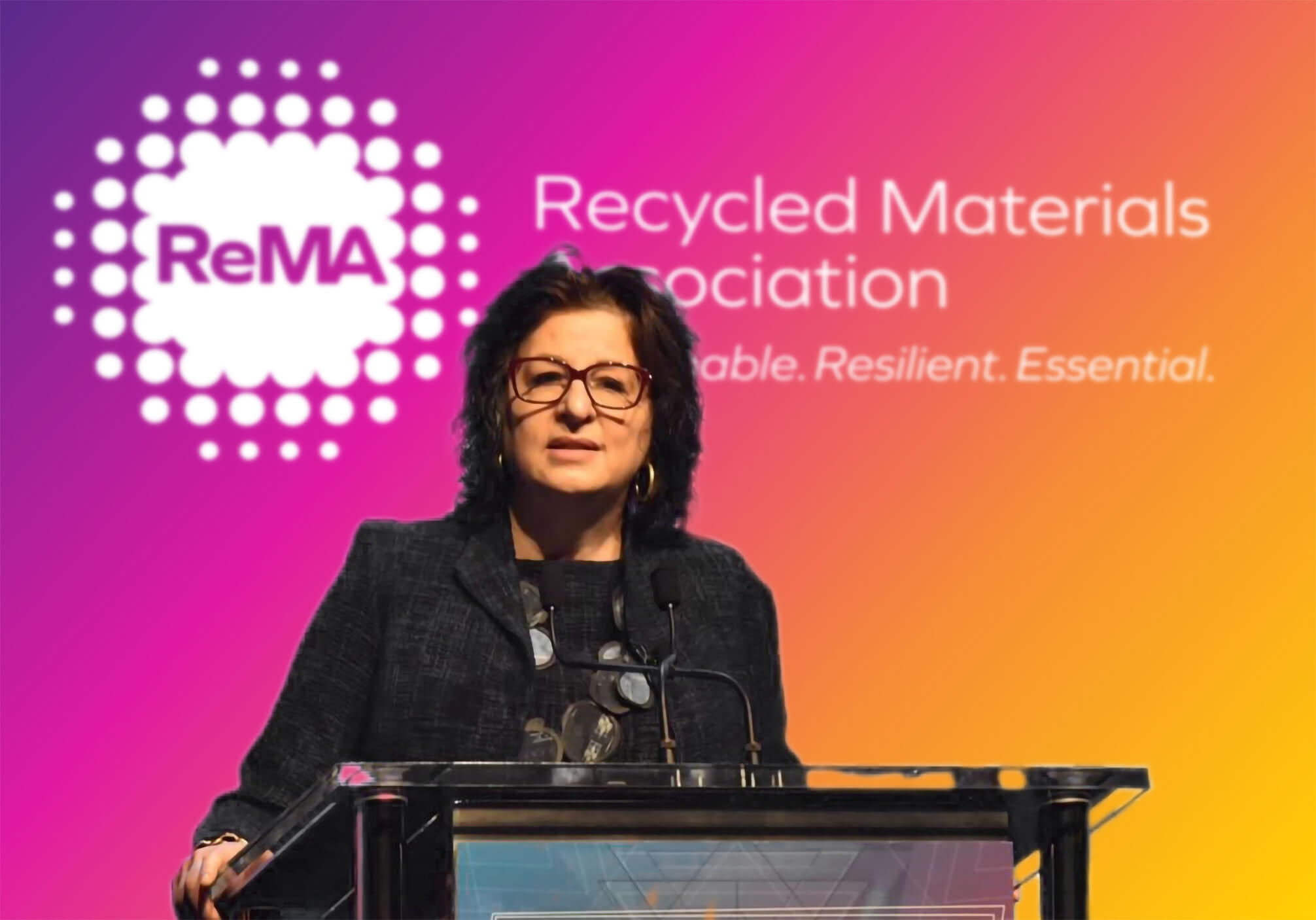 A woman in a dark blazer and glasses speaks at a podium during a Recycled Materials Association (ReMA) event, with a colorful gradient backdrop featuring the organization's logo and slogan