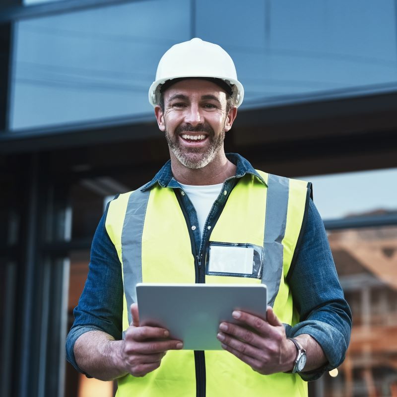 A man standing outside at a construction site with hard hat and safety vest, with a tablet in his hands smiling at the camera